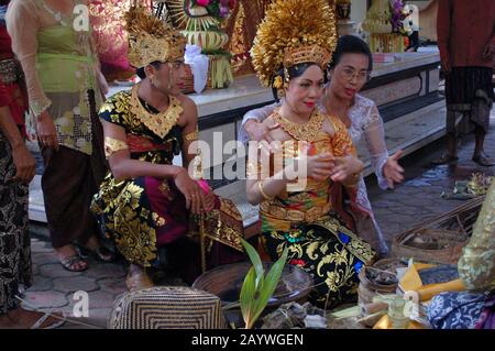 Una sposa e uno sposo Balinesi sono guidati attraverso una tradizionale cerimonia nuziale da una donna Balinese adulta. Foto Stock