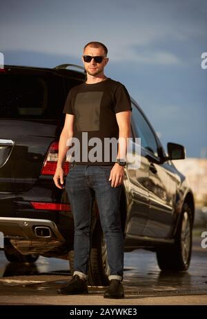 Ritratto di un uomo di successo che indossa occhiali da sole, T-shirt nera, jeans blu scuro e sneakers nere sta accanto alla sua auto nera, a tutta lunghezza Foto Stock