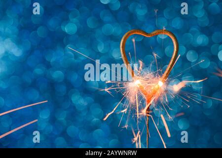 Sparkler in una forma di calore dorato che brucia su sfondo blu bokeh con copia-spazio. Matrimonio o concetto di carta d'invito di San Valentino. Foto Stock