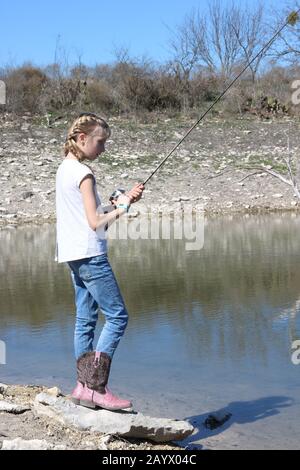 Giovane ragazza bionda con trecce francesi e stivali cowboy pesca nel lago Foto Stock