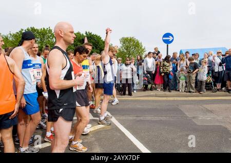 Corridori e spettatori che si raccolgono in strada pochi minuti prima dell'inizio di una corsa di beneficenza 10KM. Uomo in mezzo che si allunga prima della gara. Foto Stock