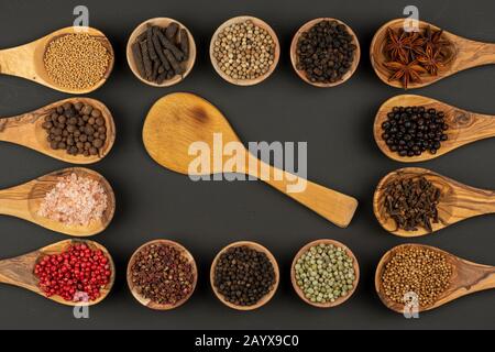 Sei piccole ciotole in legno e otto cucchiai da cucina in legno di ulivo riempiti di varie spezie e un singolo cucchiaio da cucina in legno si trovano su un grigro nero Foto Stock