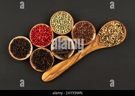Un cucchiaio da cucina in legno d'oliva e sei piccole ciotole di legno riempite con varie bucce su sfondo nero Foto Stock