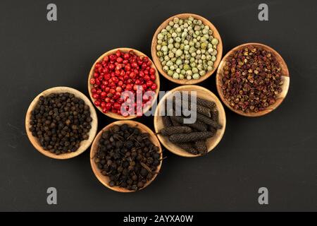 Sei piccole ciotole di legno riempite con vari grani di pepe su uno sfondo nero Foto Stock
