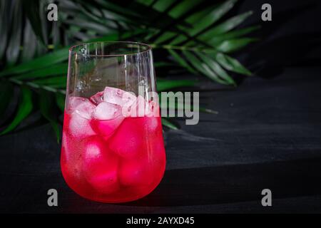 Bevanda al lampone-mirtillo in un vetro trasparente con ghiaccio. La bevanda viene versata in un bicchiere. Aggiunti rami di palma e lamponi. Sfondo nero. Cop Foto Stock