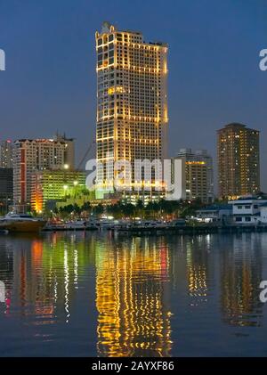 Scenario notturno di barche ormeggiate nella Baia di Manila vicino allo Yacht Club di Manila con edifici illuminati grattacieli sullo sfondo che si riflettono nell'acqua Foto Stock