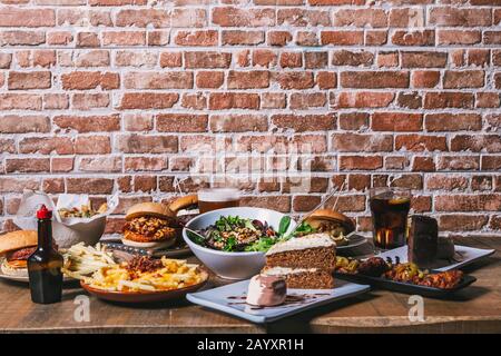 Vista sul tavolo con una varietà di piatti, hamburger, patatine fritte e insalata, bevande, ali di pollo, salsa, torta e dessert sul tavolo in legno. Restaura Foto Stock