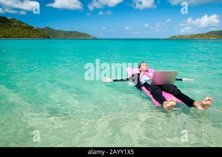 Lavoratore di ufficio in vacanza galleggiante con il suo computer portatile su una zattera gonfiabile rosa in un luminoso mare tropicale Foto Stock