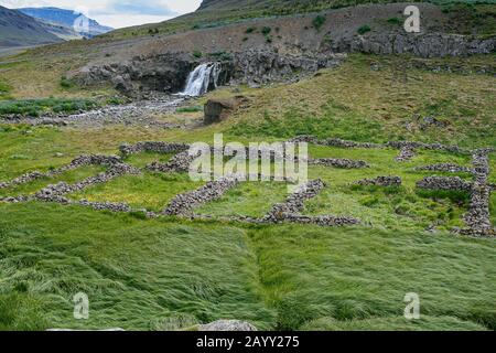 Vecchie fondamenta in pietra di ciottoli di edifici coltivati con erba verde lussureggiante con una cascata sullo sfondo Foto Stock