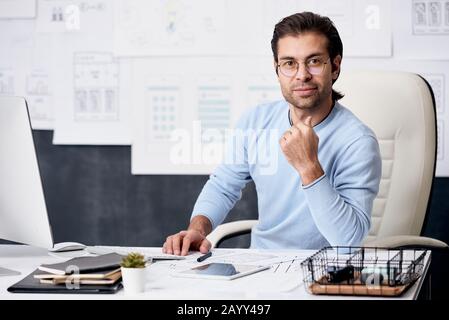 ritratto orizzontale di un giovane moderno che indossa un maglione azzurro e occhiali seduti sul tavolo dell'ufficio guardando la macchina fotografica Foto Stock