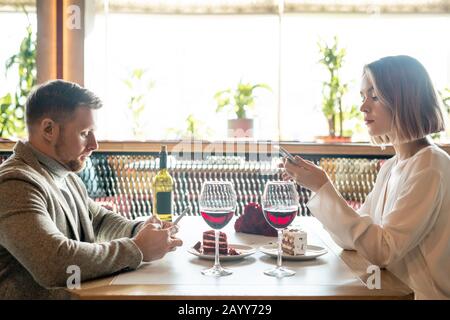 Giovane caucasico uomo e donna seduti di fronte l'uno all'altro in ristorante navigare su Internet sui loro smartphone invece di parlare l'uno con l'altro Foto Stock