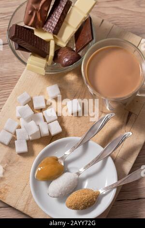 Diversi tipi di zuccheri e dolci si trovano su un banco di legno. Il miele, lo zucchero bianco sciolto e lo zucchero di canna si trovano su cucchiaini in un piattino bianco. Tea con mil Foto Stock