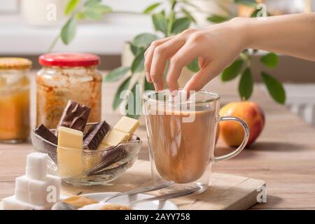 La mano del ragazzo sta tenendo un cubo di zucchero sopra un bicchiere di tè o caffè con latte. Sul tavolo si trovano diversi tipi di dolci e zucchero. Foto Stock