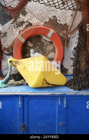 Collezione di attrezzature da pesca dai colori vivaci sul petto di legno blu con muro di pietra. L'ingranaggio include peso, anello salvavita, reti e corda. Foto Stock