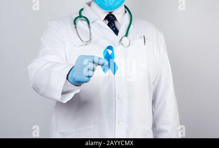 medico maschile in un cappotto bianco e cravatta si alza e tiene un nastro di seta blu in un anello, indossando guanti medici sterili blu, simbolo della lotta e del trattamento Foto Stock