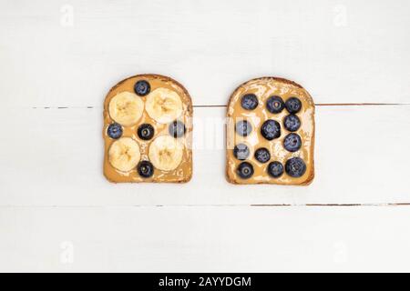 Sopra vista di pane tostato al burro di arachidi con mirtillo e banana su tavola di legno bianco. Colazione sana Foto Stock