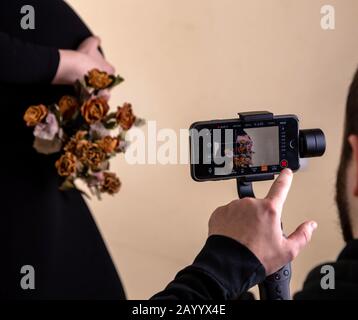 Il tipo spara un video sul telefono ad una ragazza incinta in un abito nero che posa e tiene un bouquet di rose secche nelle sue mani Foto Stock