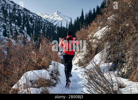 Vecchio uomo con barba grigia e giacca rossa sta correndo vicino sul sentiero in montagna in inverno. Skyrunning e trailrunning per attività all'aperto. Foto Stock