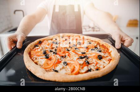 Pizza italiana calda fresca su foglio da forno, chef in grembiule tiene in mano, luce del sole Foto Stock