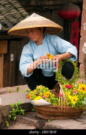 Cina: Donna Tujia che vende tiaras di fiori alla Torre porta Nord, Fenghuang, Provincia di Hunan. Fenghuang è cinese per Phoenix e si riferisce al mitico uccello da fuoco sacro che si può trovare nelle mitologie dei Persiani, Greci, Romani, Egiziani, Cinesi, E (secondo Sanchuniathon) i Fenici. La leggenda suggerisce che due phoenixes sulla scoperta della città hovered sopra per un certo tempo considerevole prima di volare riluttante via. Fenghuang è un'antica città ben conservata che risale al 248 a.C. Ospita le minoranze Miao e Tujia. Foto Stock