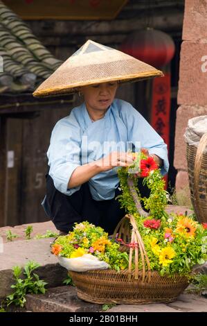 Cina: Donna Tujia che vende tiaras di fiori alla Torre porta Nord, Fenghuang, Provincia di Hunan. Fenghuang è cinese per Phoenix e si riferisce al mitico uccello da fuoco sacro che si può trovare nelle mitologie dei Persiani, Greci, Romani, Egiziani, Cinesi, E (secondo Sanchuniathon) i Fenici. La leggenda suggerisce che due phoenixes sulla scoperta della città hovered sopra per un certo tempo considerevole prima di volare riluttante via. Fenghuang è un'antica città ben conservata che risale al 248 a.C. Ospita le minoranze Miao e Tujia. Foto Stock