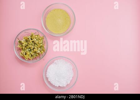 Camomilla secca, argilla gialla e sale marino in una ciotola di vetro su sfondo rosa pastello con copyspace. Il concetto di creare cosmetici naturali e ho Foto Stock