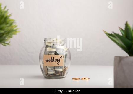 Risparmi in vaso di vetro della banca e Anelli di nozze sopra grigio Foto Stock