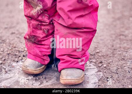 Sezione bassa di una bambina di 4 anni in pantaloni invernali rosa sporco fangoso in piedi su una pozza ghiacciata su una strada di ghiaia. Visto in Germania nel mese di febbraio. Foto Stock