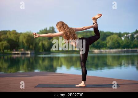 Donna sportiva in abbigliamento sportivo nero che fa yoga al lago della città. Parco verde sullo sfondo. Esercizio Natarajasana, posa del Signore della Danza, vista laterale, lunghezza intera Foto Stock