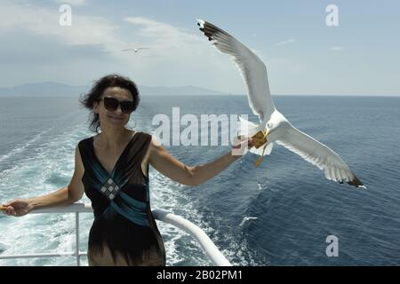 Donna che alimenta gabbiani su un traghetto da Kavala a Thassos. I gabbiani volano attraverso il cielo per prendere il cibo dalle mani dei passeggeri. Mar Egeo, Grecia Foto Stock