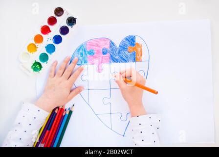 Un bambino sta disegnando cuore colorato con puzzle jigsaw su sfondo bianco come un simbolo di autismo, con matite colorate e acquerelli. Foto Stock