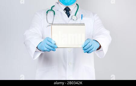 il medico maschio in un cappotto bianco e i guanti di colore blu medico tiene un taccuino con fogli bianchi bianchi bianchi bianchi bianchi bianchi su uno sfondo bianco, luogo per un'iscrizione Foto Stock