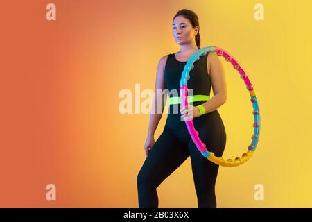 Giovane caucasico più dimensione femmina modello di formazione su gradiente arancio-pesca sfondo in luce al neon. In posa sicura con hula hoop. Concetto di sport, stile di vita sano, corpo positivo, uguaglianza. Foto Stock