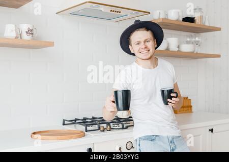 Giovane uomo sorride e condivide caffè preparato di fresco in tazza nera ceramica. Luminoso sfondo cucina Foto Stock