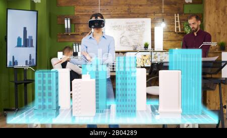 Architect Woman WHOUWER VR Headset sta guardando proiezioni olografiche della costruzione di una città. Riorganizza i piani di una torre aziendale Foto Stock