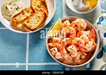Scampi speziati freschi o aragosta in una ciotola con un cuneo di limone servito con baguette tostate e burro sul lato su un tavolo piastrellato blu in estate Foto Stock