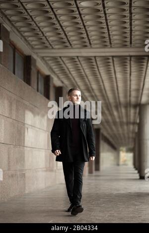 Uomo elegante in trench nero e passeggiate con il collo di polo presso l'edificio modernista Foto Stock