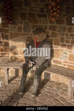 Scarecrow Vestito in vestiti di campagna Seduta su una panca di legno in una Casa estiva di pietra nel Giardino di verdure a Rosemoor in Devon rurale, Inghilterra, Regno Unito Foto Stock