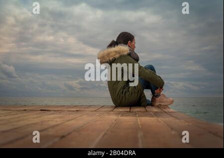 Donna che indossa un cappotto parka seduto accanto al mare, cielo tempestoso, concetto di lonelità Foto Stock