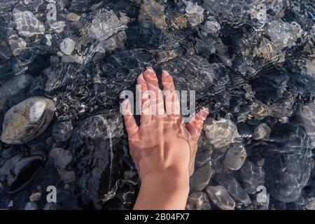 La mano di una giovane donna che tocca ciottoli sott'acqua pulita e trasparente di un fiume di montagna con luce solare che si riflette e tremolante Foto Stock