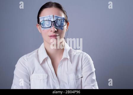 Ritratto di una giovane ragazza, dietro i cui occhiali i suoi occhi sono chiusi con dollaro fatture inserite per i bicchieri. Concetto di business sul backgroun grigio Foto Stock