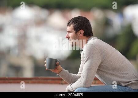Vista laterale ritratto di un uomo serio seduto su una panchina che tiene la tazza del caffè contemplando le viste in una città Foto Stock