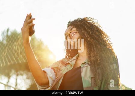 Giovane ragazza africana americana felice che usa il cellulare intelligente in città parco all'aperto - Afro girl divertirsi con le nuove reti sociali video call - Technolo Foto Stock