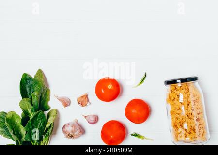 Fusilli maccheroni in vasetto di vetro accanto a pomodori freschi, foglie di aglio e spinaci, piatto su fondo bianco con spazio di copiatura. Foto Stock