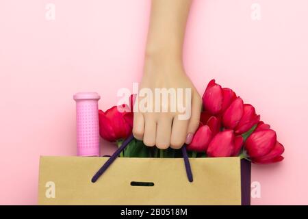 Concetto del regalo con vino e tulipani rossi nel sacchetto di carta sullo sfondo rosa. Disposizione piatta, vista dall'alto, spazio di copia. La mano della donna tiene un presente a. Foto Stock