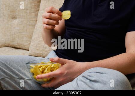 Un uomo seduto su un divano che mangia patatine da un piatto di vetro. Primo piano. Foto Stock