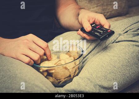 Un uomo si siede su un divano che mangia patatine dalla vetreria e cambia i canali con un telecomando TV. Tonato. Primo piano. Foto Stock