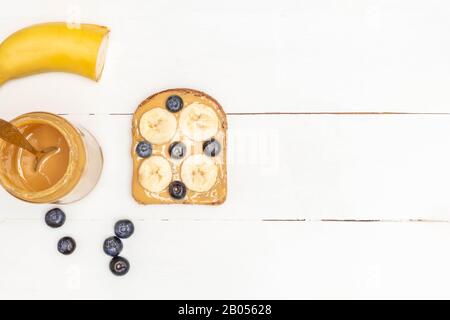 Colazione toast. Toast al burro di arachidi con mirtillo e banana su tavola di legno bianco. Immagine con spazio di copia, vista dall'alto Foto Stock