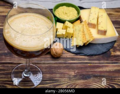 Versare la birra rossa dell'abbazia belga in vetro e piatto di pietra con formaggi prodotti da monaci di latte vaccino con fenugreek e semi di senape vicino Foto Stock