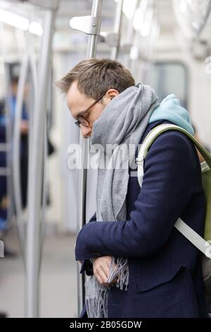 Uomo stanco in occhiali in metro treno. Giovane esausto maschio in piedi e dormire in metropolitana dopo il lavoro, soft focus. Affaticamento cronico, stanchezza, sonno Foto Stock